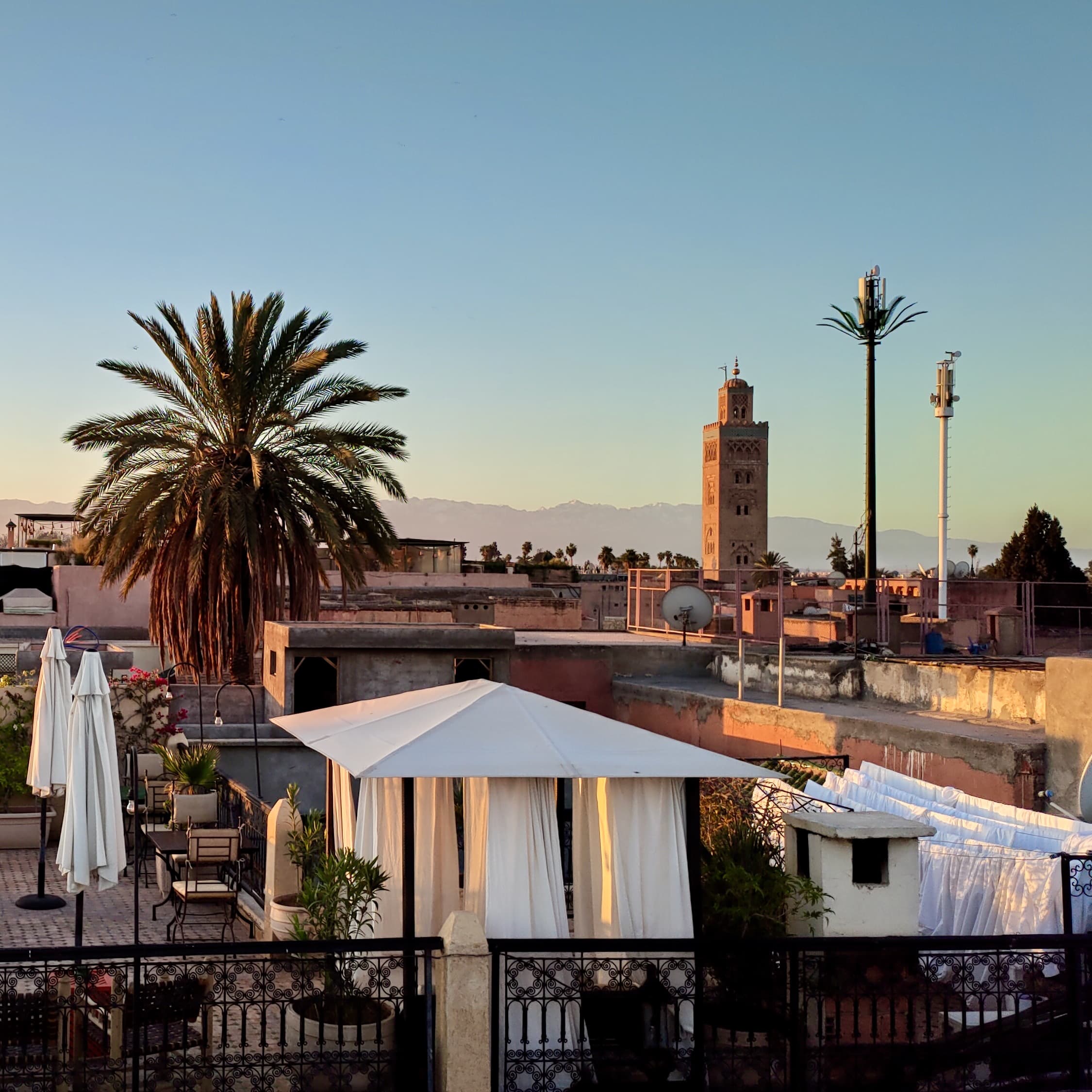 The sunset view from the terrace, with golden light illuminating one side of the Khoutoubia mosque. The Atlas Mountains are visible on the horizon.